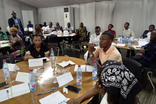 Students from Prowibo's Guinea programme sitting around tables in a conference hall. One male student is speaking into a microphone.