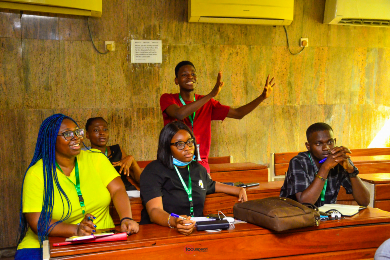 Five Nigerian students sitting in a classroom. One of the students is standing out while he speaks. The others are listening intently.