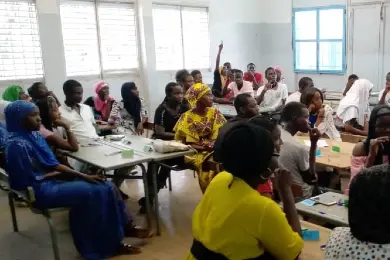 A class of Senegalese students sitting in a classroom facing the teacher, who is out of the shot. One student at the back of the class has their arm raised.